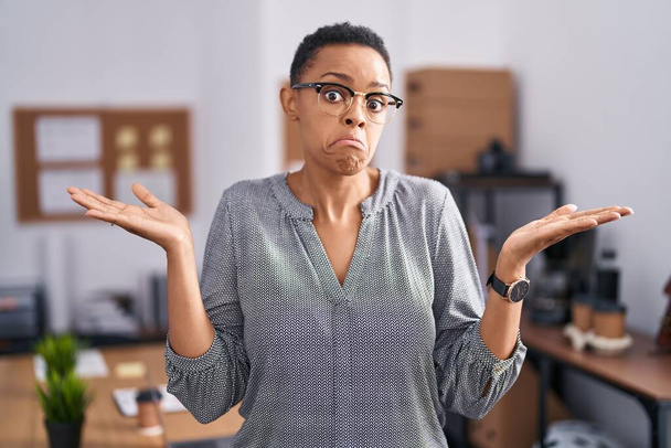オフィスで働くアフリカ系アメリカ人女性は、眼鏡をかけずに腕や手を上げて混乱した表情をしていました。疑わしい概念.  - 写真・画像