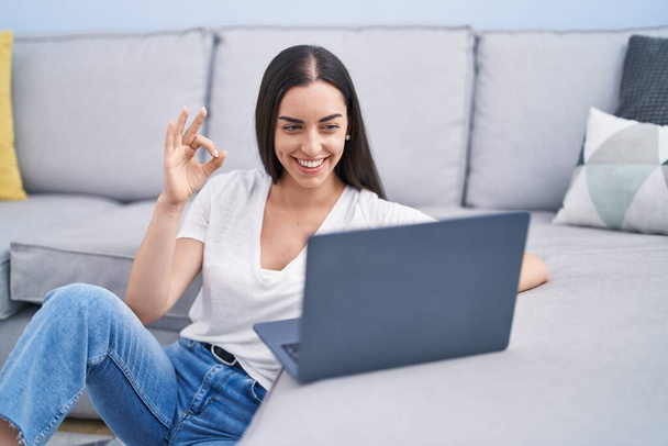 Mujer morena joven usando el ordenador portátil en casa haciendo signo de ok con los dedos, sonriendo gesto amistoso excelente símbolo  - Foto, Imagen
