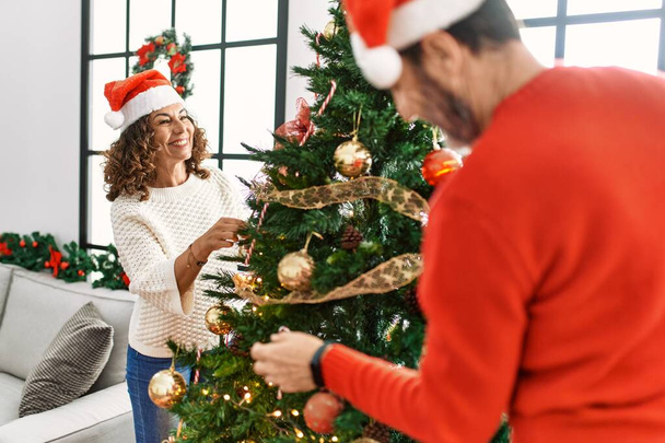 Pareja hispana de mediana edad sonriendo feliz decorando el árbol de Navidad en casa. - Foto, imagen