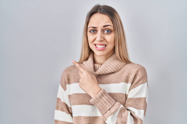 Giovane donna bionda indossa maglione dolcevita su sfondo isolato indicando da parte preoccupato e nervoso con indice, preoccupato e sorpreso espressione  - Foto, immagini