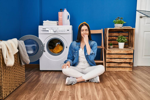 Hispanic woman doing laundry sitting on the floor looking stressed and nervous with hands on mouth biting nails. anxiety problem.  - Fotó, kép