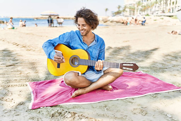Junger hispanischer Mann spielt klassische Gitarre und sitzt am Strand auf Sand. - Foto, Bild