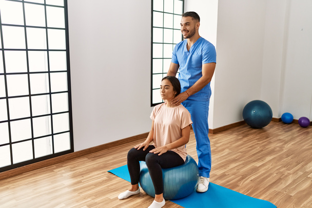 Hombre y mujer latinos vistiendo uniforme de fisioterapeuta usando el cuello de masaje de bola en forma en el centro de rehabilitación - Foto, imagen