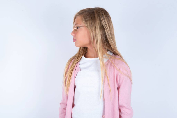 Blondes kleines Mädchen mit rosa Jacke und Brille vor weißem Hintergrund, das zur Seite schaut - Foto, Bild