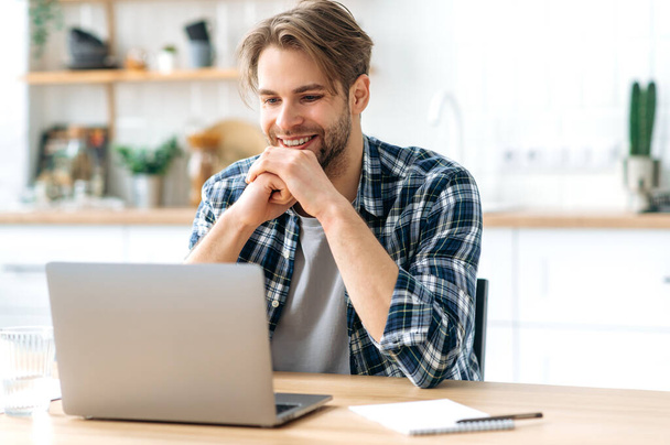 Positive successful modern caucasian male freelancer, programmer, stylishly dressed, sits at desk with laptop in a kitchen at home, working on a creative project, looking to the side, smiling happily - Photo, Image