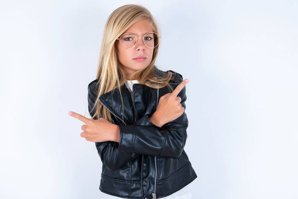 Serious beautiful blonde little girl wearing biker jacket and glasses over white background crosses hands and points at different sides hesitates between two items. Hard decision concept - Photo, Image