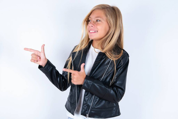 Optimistisch schönes blondes kleines Mädchen in Bikerjacke und Brille über weißem Hintergrund, das mit beiden Händen auf den leeren Raum zeigt. - Foto, Bild