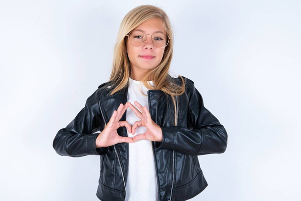 Serious beautiful blonde little girl wearing biker jacket and glasses over white background keep hands in heart shape - Photo, Image