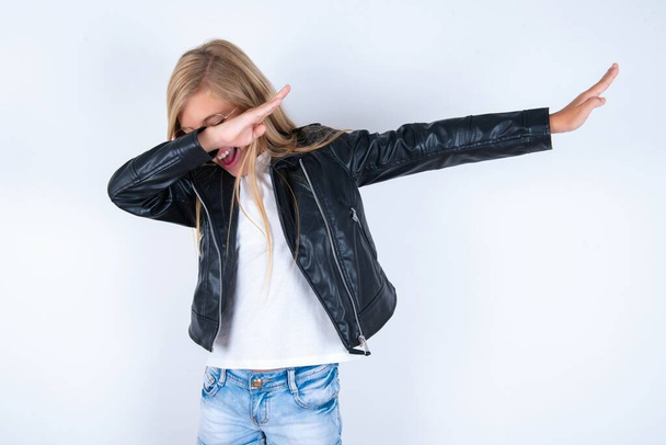 Photo of funky beautiful blonde little girl wearing biker jacket and glasses over white background show disco move dab - Foto, afbeelding