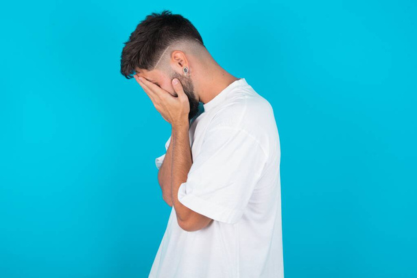 Sad bearded caucasian man wearing white T-shirt over blue background covering face with hands and crying. - Foto, Imagen
