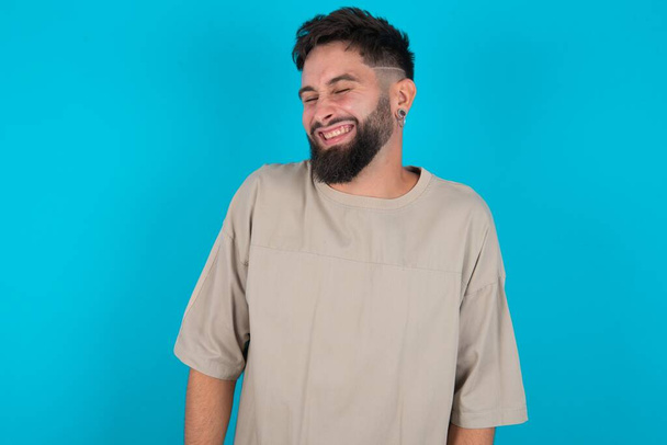 Positieve bebaarde blanke man draagt casual T-shirt over blauwe achtergrond met overblijde uitdrukking sluit de ogen en lacht toont witte perfecte tanden - Foto, afbeelding