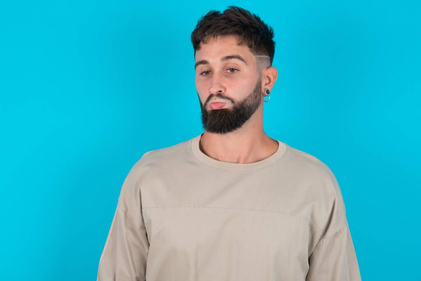 Недовольный бородатый кавказский мужчина в повседневной футболке на синем фоне сумочки губы и имеет несчастливое выражение лица смотрит в сторону стоит обижен. Депрессивная разочарованная модель. - Фото, изображение