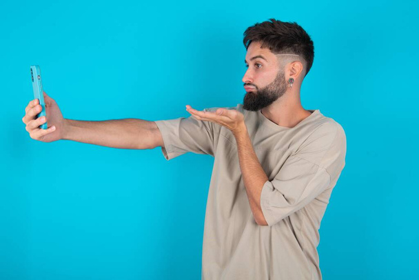 barbudo caucásico hombre usando casual camiseta sobre fondo azul sopla aire beso en la cámara de teléfono inteligente y toma selfie, envía mwah a través de llamada en línea. - Foto, imagen