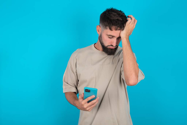 Upset depressed bearded caucasian man wearing casual T-shirt over blue background makes face palm as forgot about something important holds mobile phone expresses sorrow and regret blames - Fotó, kép