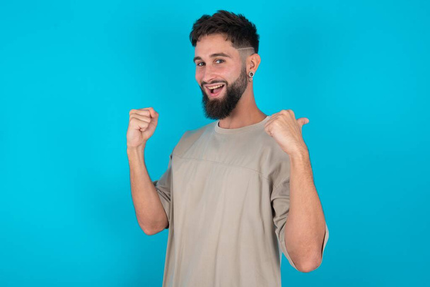 Hooray and cool bearded caucasian man wearing casual T-shirt over blue background point back empty space hand fist - Photo, Image