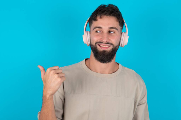 bebaarde blanke man draagt casual T-shirt over blauwe achtergrond luistert audio track via draadloze hoofdtelefoon punten duim weg adverteert kopieerruimte - Foto, afbeelding
