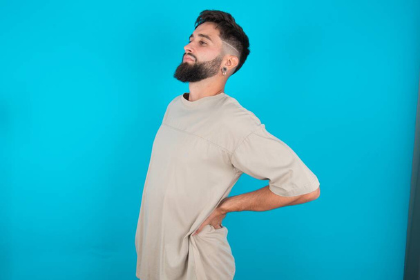 barbuto caucasico uomo indossa casual t-shirt su sfondo blu ottenuto mal di schiena - Foto, immagini