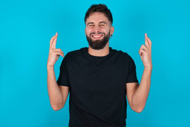 Jonge latino bebaarde man met een zwart T-shirt tegen een blauwe achtergrond heeft grote hoop, kruist vingers, gelooft in geluk, glimlacht breed. Mensen en wensconcept - Foto, afbeelding