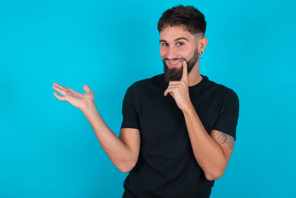 drôle hispanique barbu homme portant noir T-shirt debout sur fond bleu tenir palme ouverte nouveau produit grande proposition - Photo, image