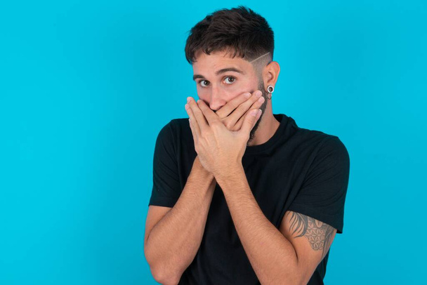 Young hispanic bearded man wearing black T-shirt standing against blue background shocked covering mouth with hands for mistake. Secret concept. - Photo, Image