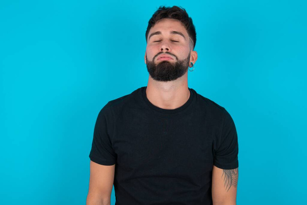 Young hispanic bearded man wearing black T-shirt standing against blue background looking sleepy and tired, exhausted for fatigue and hangover, lazy eyes in the morning. - Foto, Bild