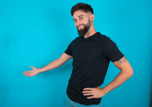Joven hombre barbudo hispano vistiendo camiseta negra de pie sobre fondo azul sintiéndose feliz y alegre, sonriendo y dándote la bienvenida, invitándote a entrar con un gesto amistoso - Foto, imagen