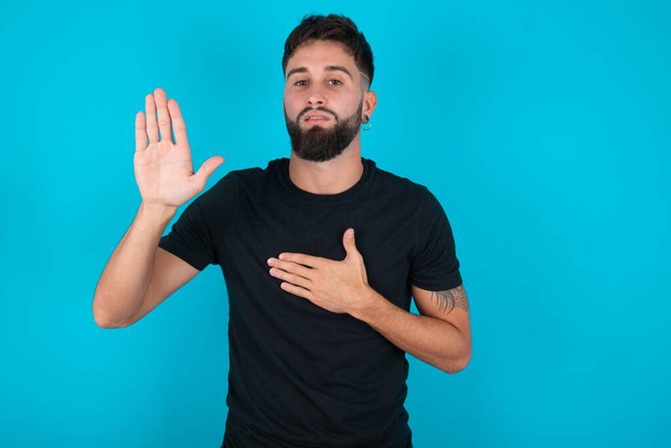 Giovane uomo barbuto ispanico indossa una t-shirt nera in piedi su sfondo blu giurando con mano sul petto e palmo aperto, facendo un giuramento di fedeltà - Foto, immagini