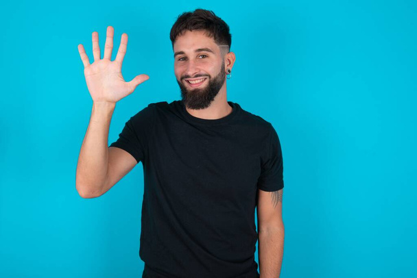 Young hispanic bearded man wearing black T-shirt standing against blue background showing and pointing up with fingers number five while smiling confident and happy. - Photo, Image