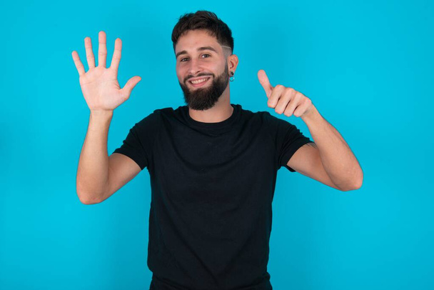 Молодий іспаномовний бородатий чоловік у чорній футболці стоїть на синьому фоні, показуючи і вказуючи пальцями номер шість, посміхаючись впевнено і щасливо. - Фото, зображення