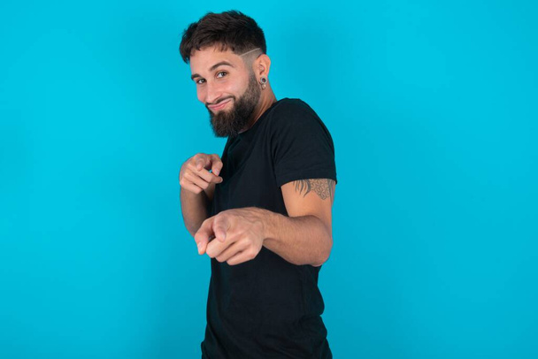 Joyful hispanic bearded man wearing black T-shirt standing against blue background wink and points index fingers at camera, chooses someone, makes finger gun pistol. - Photo, Image