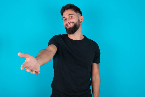 homme barbu hispanique portant T-shirt noir debout sur fond bleu souriant amical offrant poignée de main comme salutation et accueil. Une entreprise prospère. - Photo, image