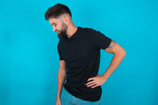 hombre barbudo hispano vistiendo camiseta negra de pie sobre fondo azul Sufrimiento de dolor de espalda, tocando la espalda con la mano, dolor muscular - Foto, Imagen