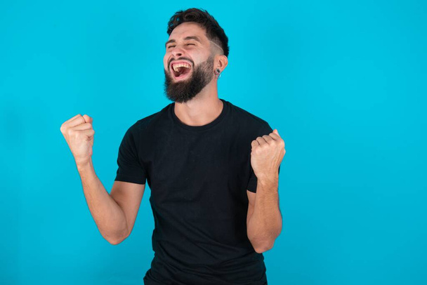 homme barbu hispanique portant T-shirt noir debout sur fond bleu célébrant surpris et étonné pour le succès avec les bras levés et les yeux fermés. Concept gagnant. - Photo, image