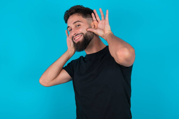 hispanique barbu homme portant noir T-shirt debout sur fond bleu Essayant d'entendre les deux mains sur le geste de l'oreille, curieux de commérages. Problème auditif, sourd - Photo, image