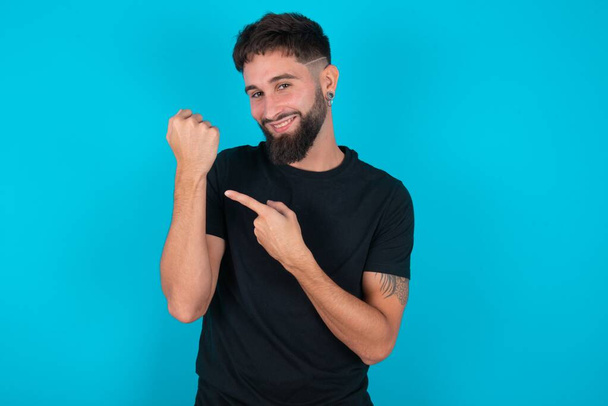hispanique barbu homme portant noir T-shirt debout sur fond bleu Dans la hâte pointant vers montre-bracelet, impatience, regarder la caméra avec une expression détendue - Photo, image