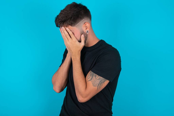 Sad hispanic bearded man wearing black T-shirt standing against blue background crying covering her face with her hands. - Photo, Image