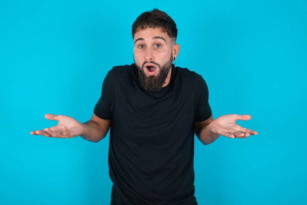 Frustrated hispanic bearded man wearing black T-shirt standing against blue background feels puzzled and hesitant, shrugs shoulders in bewilderment, keeps mouth widely opened, doesn't know what to do. - Photo, Image