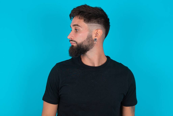 Foto de perfil lateral de un hombre barbudo hispano vestido con una camiseta negra de pie sobre fondo azul sin sonreír atento escuchar concentrado - Foto, imagen