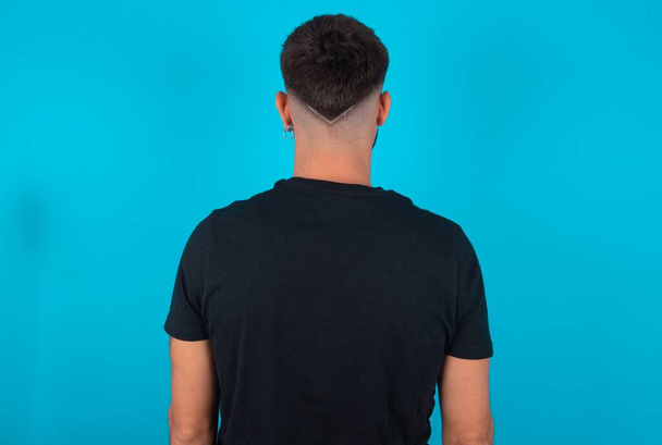 La vue arrière d'un homme barbu hispanique portant un T-shirt noir debout sur fond bleu. Studio Shoot. - Photo, image