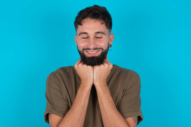 Веселый молодой латиноамериканец, бородатый мужчина, стоящий на синем фоне, робко удовлетворен выражением лица, широко улыбается, показывает белые зубы, эмоции людей - Фото, изображение