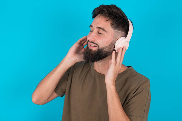 young hispanic bearded man standing against blue background with headphones on her head, listens to music, enjoying favourite song with closed eyes, holding hands on headset. - Photo, Image