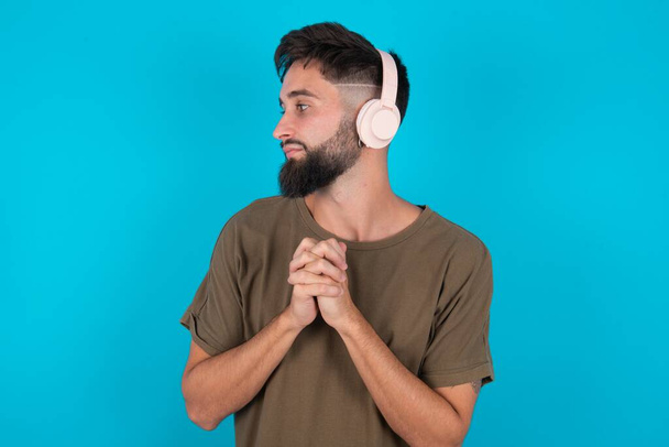młody latynoski brodaty mężczyzna stojący na niebieskim tle nosi słuchawki stereo słuchając muzyki skoncentrowanej i patrząc na bok z zainteresowaniem. - Zdjęcie, obraz