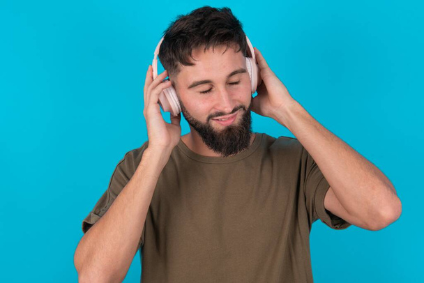молодий іспаномовний бородатий чоловік, що стоїть на синьому фоні, носить стерео навушники, слухає музику, зосереджену вбік. Люди хобі концепція способу життя - Фото, зображення