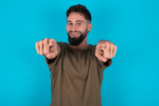joven hombre barbudo hispano de pie sobre fondo azul alegre y sonriente apuntando a la cámara - Foto, Imagen