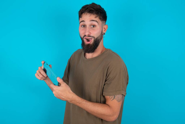 Buen adicto alegre joven hispano barbudo hombre de pie sobre fondo azul utilizando gadget juego de red - Foto, imagen