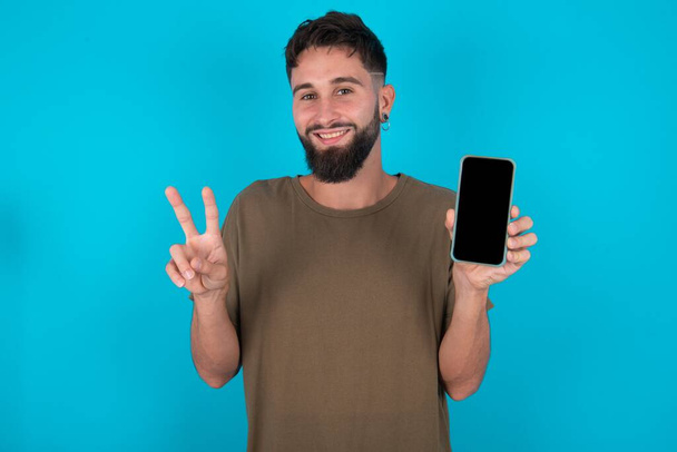 young hispanic bearded man standing against blue background holding modern device showing v-sign - Foto, imagen
