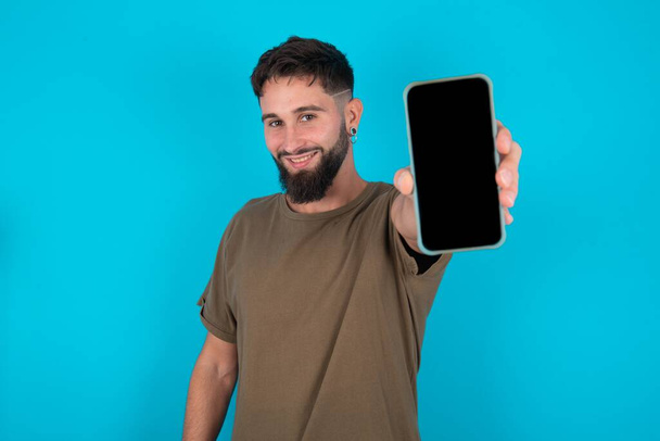 Encantador joven barbudo hispano adorable de pie sobre fondo azul que sostiene el dispositivo moderno, mostrando el teléfono inteligente de pantalla negra - Foto, Imagen