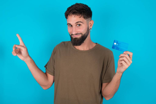 Χαμογελώντας νεαρός ισπανόφωνος γενειοφόρος άνδρας στέκεται πάνω σε μπλε φόντο δείχνει χρεωστική κάρτα δείχνοντας κενό χώρο δάχτυλο - Φωτογραφία, εικόνα