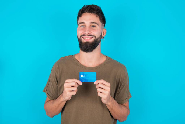 青い背景に立っている若いヒスパニックひげの男の写真 ポジティブな笑顔 クレジットカードの所得の給与を保持します. - 写真・画像