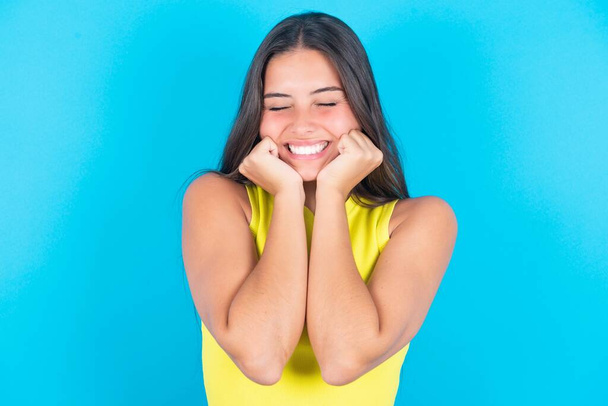 Portrait of beautiful brunette woman wearing yellow tank top over blue background being overwhelmed, expressing excitement and happiness with closed eyes and hands near face. - Foto, imagen